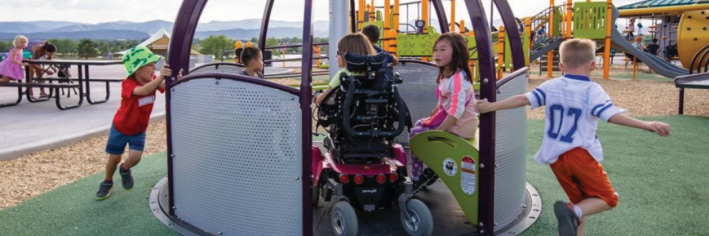 Des enfants jouent sur une aire de jeux accessible par une journée d'été ensoleillée. Un enfant en fauteuil roulant se trouve sur un carrousel accessible aux fauteuils roulants avec trois autres enfants. Deux autres enfants courent et tirent le carrousel pour le faire tourner pour les enfants assis dessus. 