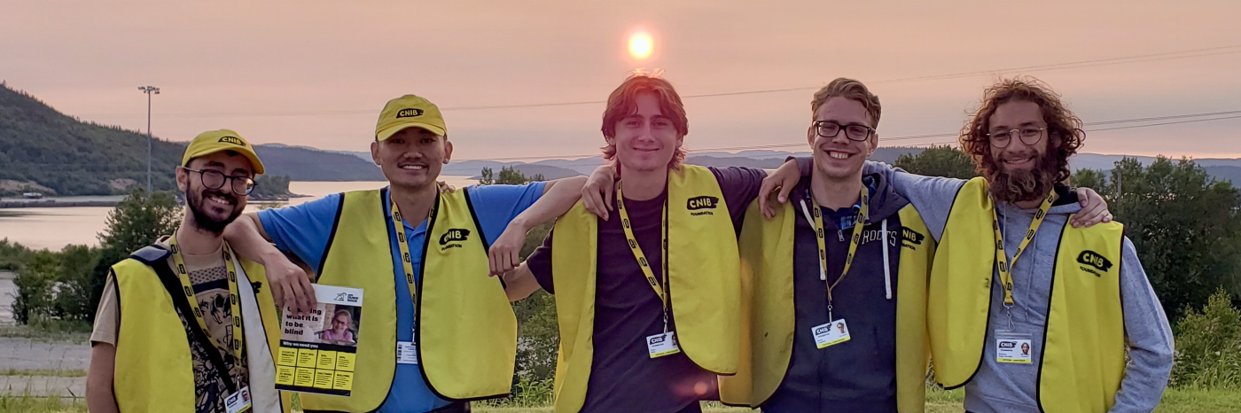 Cinq collecteurs de fonds travaillant pour le compte d'INCA se tiennent côte à côte, les bras enlacés, devant un coucher de soleil d'un rouge éclatant. Ils portent tous un gilet jaune d'INCA et un cordon avec photo d'identité. Les deux collecteurs de fonds de gauche portent également une casquette de baseball jaune d'INCA et tiennent de grandes cartes présentant des faits sur la cécité.