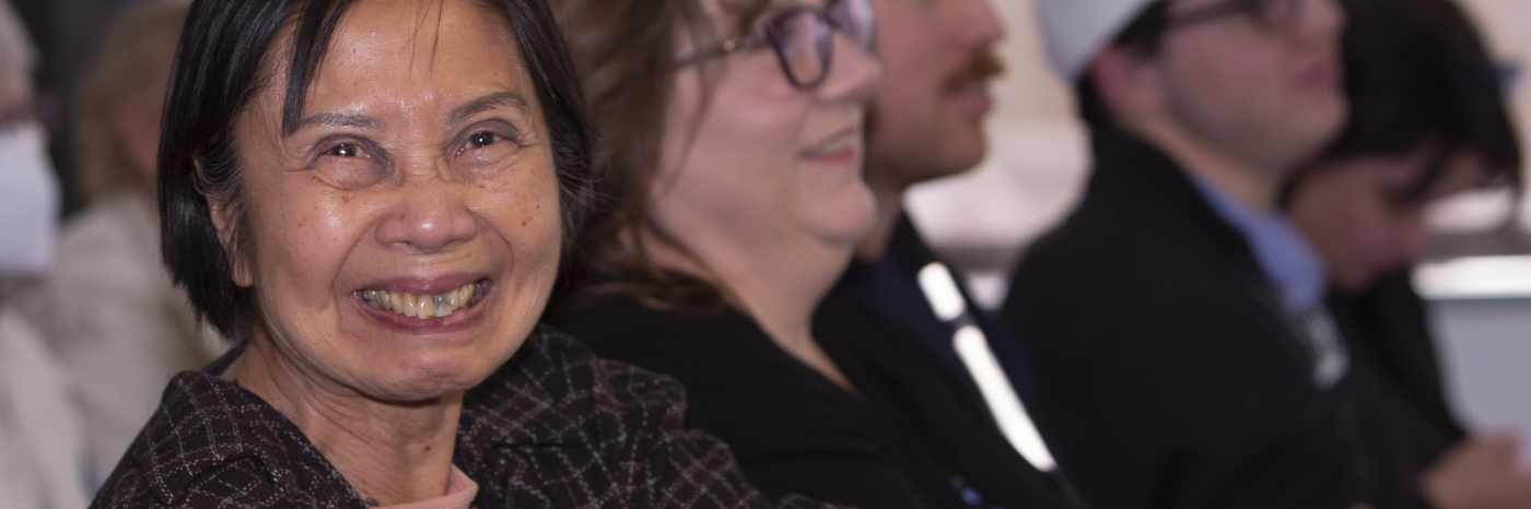 Une femme ayant une perte de vision regarde directement la caméra et sourit. Elle est assise au milieu d'un grand groupe de personnes hors champ en arrière-plan