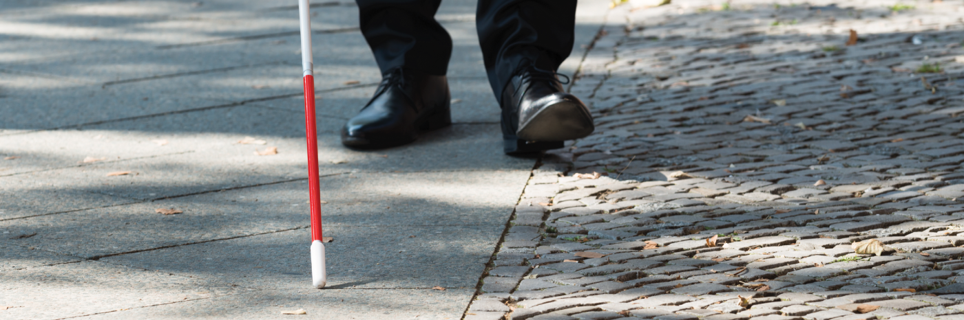 Une canne blanche balaie un trottoir. En arrière-plan, on aperçoit des chaussures d’un homme derrière la pointe de la canne.