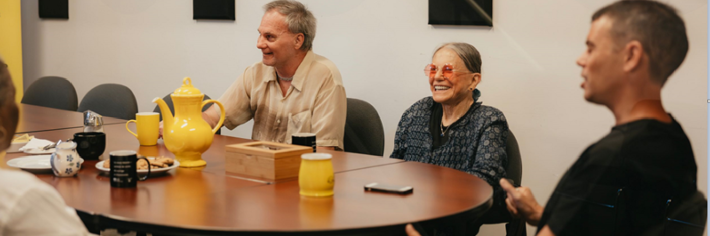 Quatre personnes sont assises autour d'une table de réunion, riant et discutant, avec une théière, des tasses et des snacks.