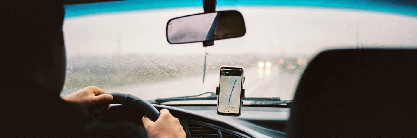 À l'intérieur d'une voiture de covoiturage, la photo est prise du point de vue du passager sur la banquette arrière. Il y a un téléphone intelligent monté sur le tableau de bord de la voiture affichant une application de rideshare. Le conducteur a les mains sur le volant et il pleut à l'extérieur. 