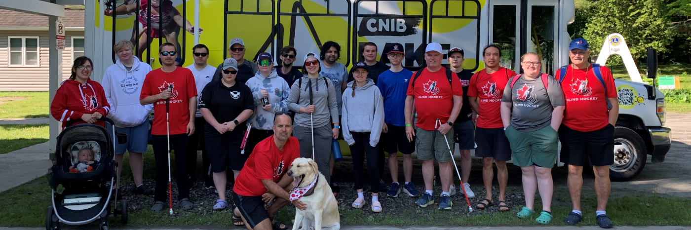 Environ 20 personnes se tiennent devant le nouveau minibus du Camp Lake Joe d'INCA. Plusieurs portent des t-shirts rouges du Hockey sonore Canada et tiennent des cannes blanches. Un chien-guide se trouve devant.