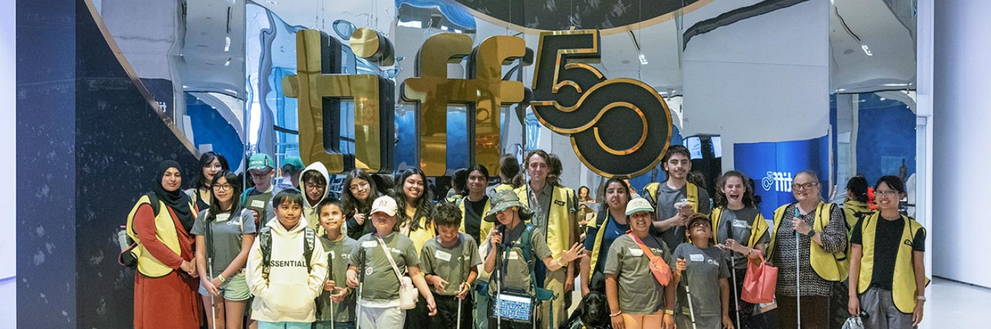 Dans le hall du TIFF Lightbox, un grand groupe de jeunes campeurs et de bénévoles d’INCA posent pour une photo de groupe devant une enseigne géante « TIFF 50 ».