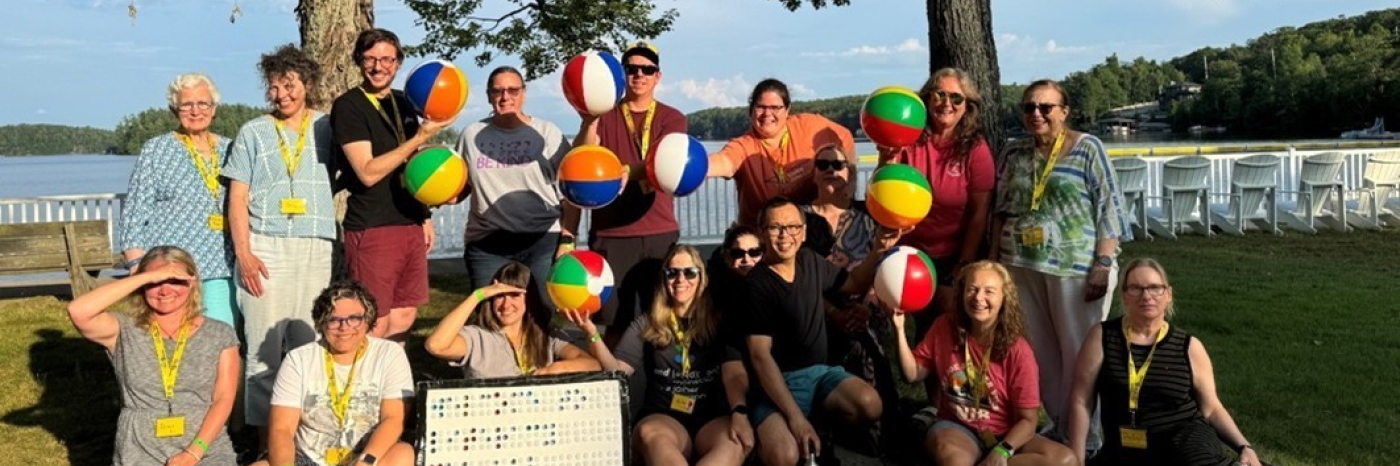 Un groupe d'employés et de bénévoles du Camp Lake Joe d’INCA souriant à l'extérieur par une journée ensoleillée au bord du lac Joseph. Ils tiennent des ballons de plage colorés sur lesquels est imprimé « BRL » pour représenter le mot braille. Ils posent sous de grands arbres avec des chaises blanches et de l'eau en arrière-plan.