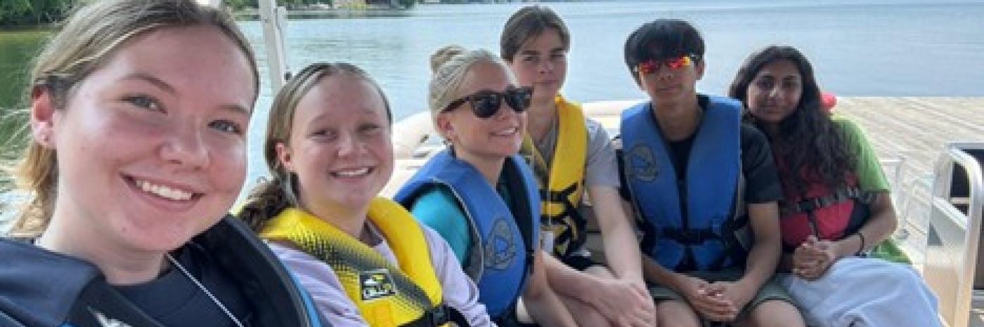 Des jeunes hommes et femmes sont sur un bateau ponton sur le lac, vêtus de gilets de sauvetage colorés.