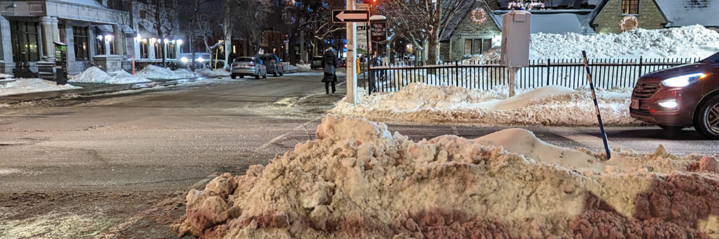 Un passage piéton dans un quartier urbain est montré un soir d'hiver. Le passage piéton est bloqué par un banc de neige de plusieurs mètres de haut.