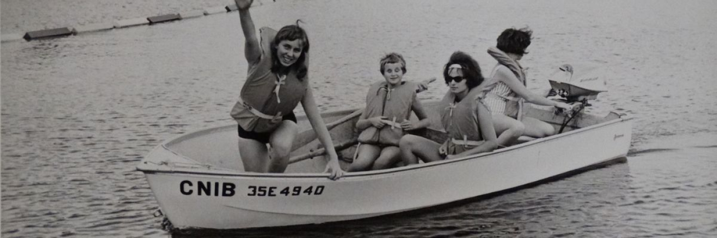 Une photo en noir et blanc montrant deux femmes et deux enfants portant des gilets de sauvetage. Ils sourient, assis dans un bateau sur lequel figure l'inscription « INCA », sur un lac.