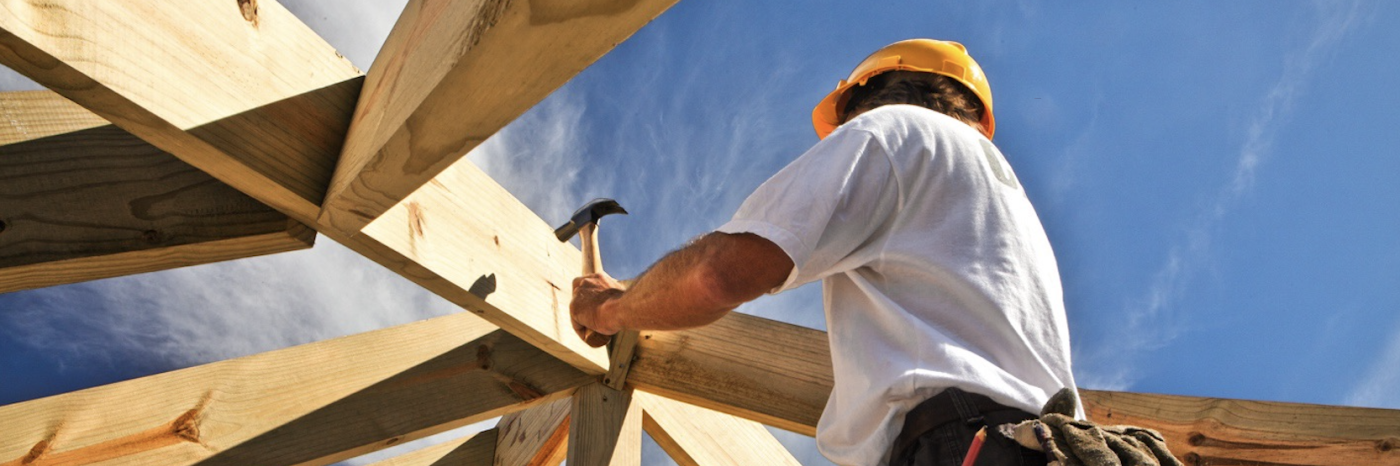 un ouvrier du bâtiment portant un casque de chantier utilise un marteau pour fixer des poutres de toit en bois, vu d'en bas sous un ciel bleu éclatant.