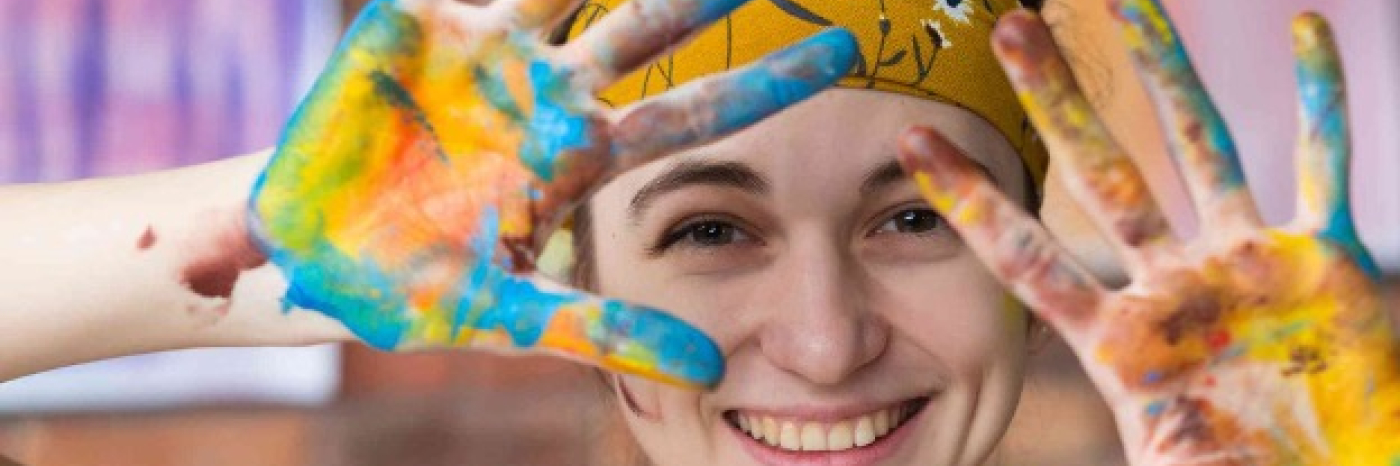 Une jeune femme sourit à la caméra. Ses cheveux sont retenus par un bandana doré à fleurs. Elle tient ses mains ouvertes devant elle, et celles-ci sont recouvertes de peinture aux couleurs vives.