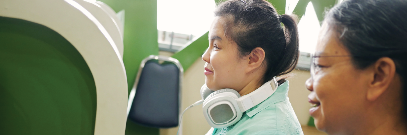 Dans un bureau cloisonné, une femme est assise devant un poste de travail informatique et tape sur un clavier. Un grand casque audio est suspendu autour de son cou. À côté d’elle, un collègue est assis à un autre poste de travail. Ils examinent un document lié au travail sur l’écran de l’ordinateur.