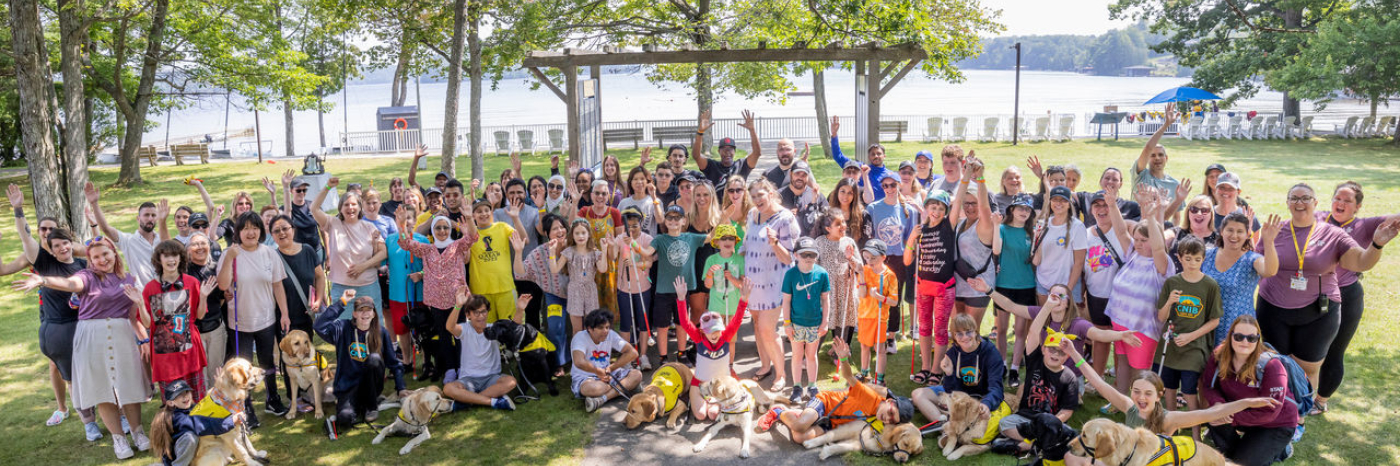 Au Camp Lake Joe, un large groupe de personnes de tous âges, dont bon nombre sont accompagnées de leurs chiens compagnons ou tiennent une canne blanche, lèvent joyeusement les mains en l'air, le lac en toile de fond.