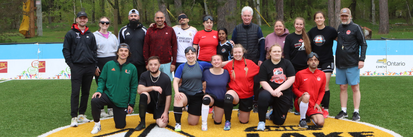 Un groupe de joueurs de soccer aveugles, de tailles, d'âges et de couleurs de peau variés en tenue de sport se tient derrière un ballon de cécifoot sur le terrain de soccer accessible du camp Lake Joe d’INCA.