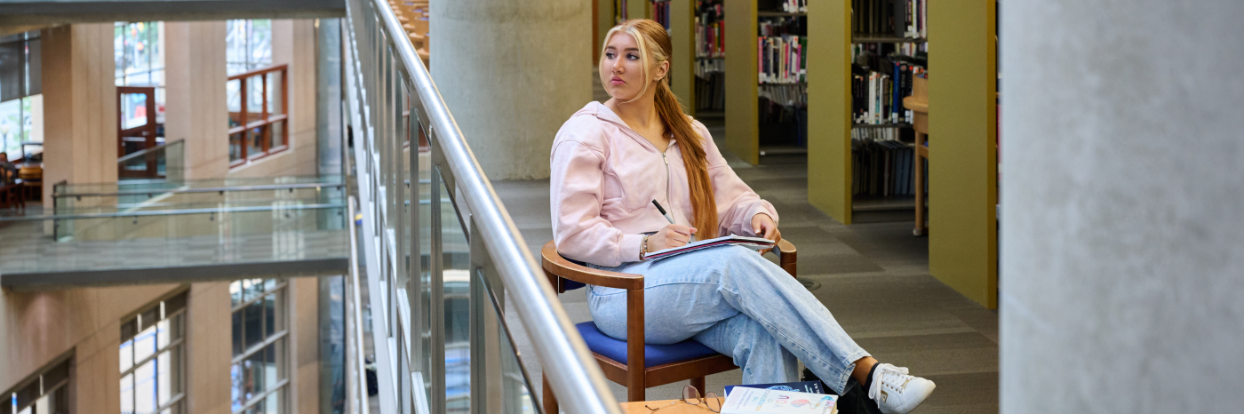 Ashlyn, vêtue d'un pull rose et d'un jean, est assise sur une chaise en bois près de la balustrade en verre d'une bibliothèque à plusieurs étages. Elle tient un cahier et un stylo sur ses genoux et regarde par-dessus le bord de la balustrade.