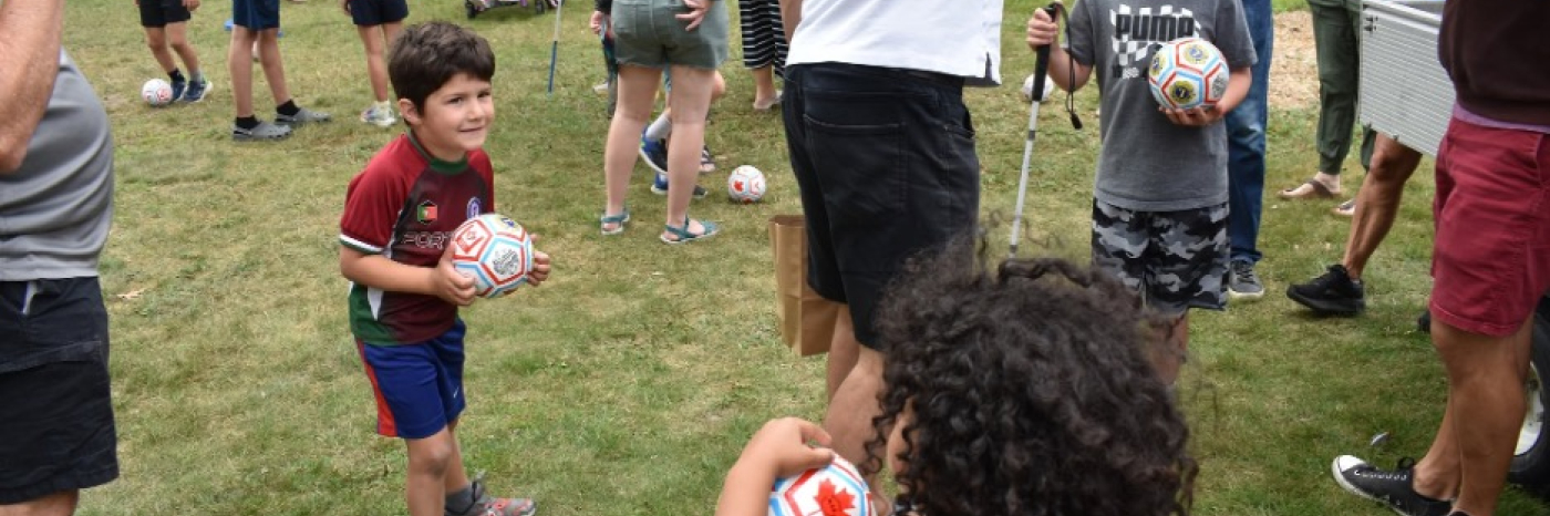 Une foule d’enfants et d’adultes se rassemble sur une pelouse pour une activité de football. Beaucoup d’entre eux tiennent de petits ballons de football blancs dans leurs mains ou les font rouler à leurs pieds.
