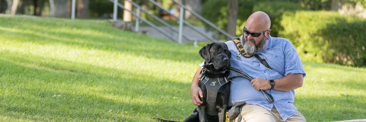 Jason est assis sur un rebord dans un parc, à côté de Midas, son chien-guide noir. Jason regarde Midas en souriant ; il porte un t-shirt bleu clair à col, un pantalon beige et des lunettes de soleil noires.