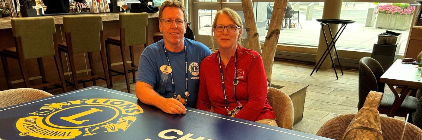 Jerry et Jenn sont assis ensemble derrière une table sur laquelle sont imprimés le logo du Lions International et les mots "Cheers to Kindness".