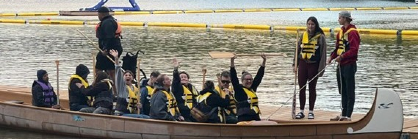 Un groupe de personnes dans un grand canoë Voyageur à quai. Elles portent des gilets de sauvetage jaunes et ont les mains levées en l'air.
