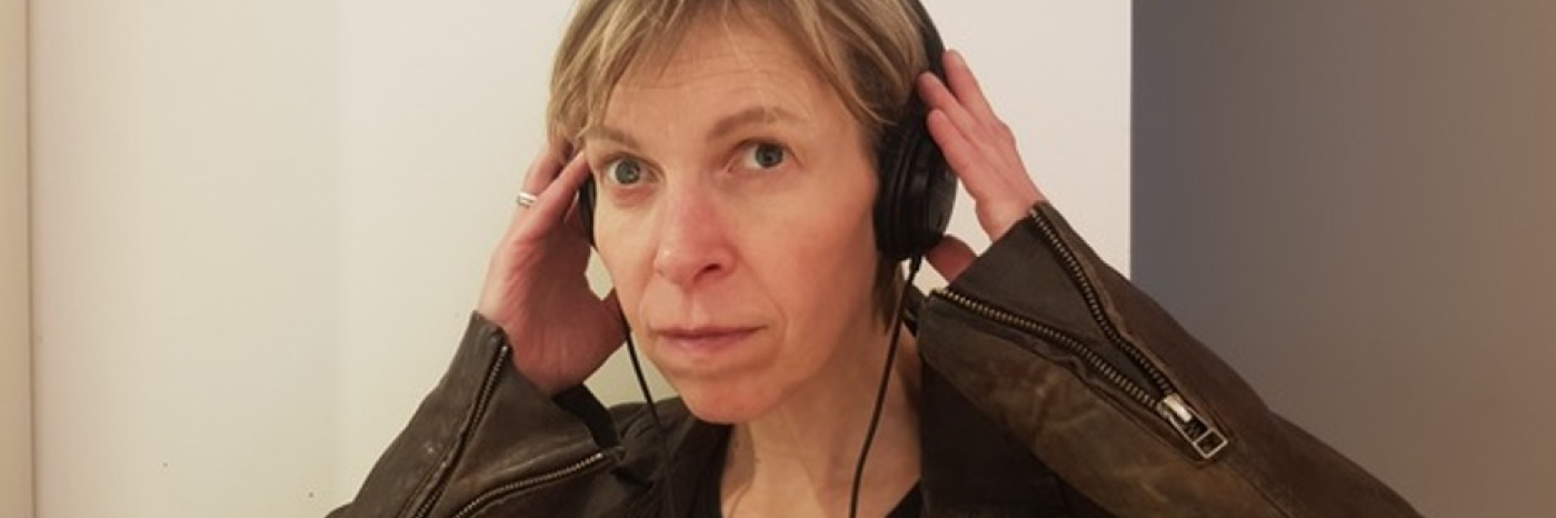 Photo en gros plan d'Alex regardant l'objectif. Elle pose ses deux mains sur le casque qu'elle porte. Elle porte une veste en cuir marron dotée de fermetures éclair aux poignets.