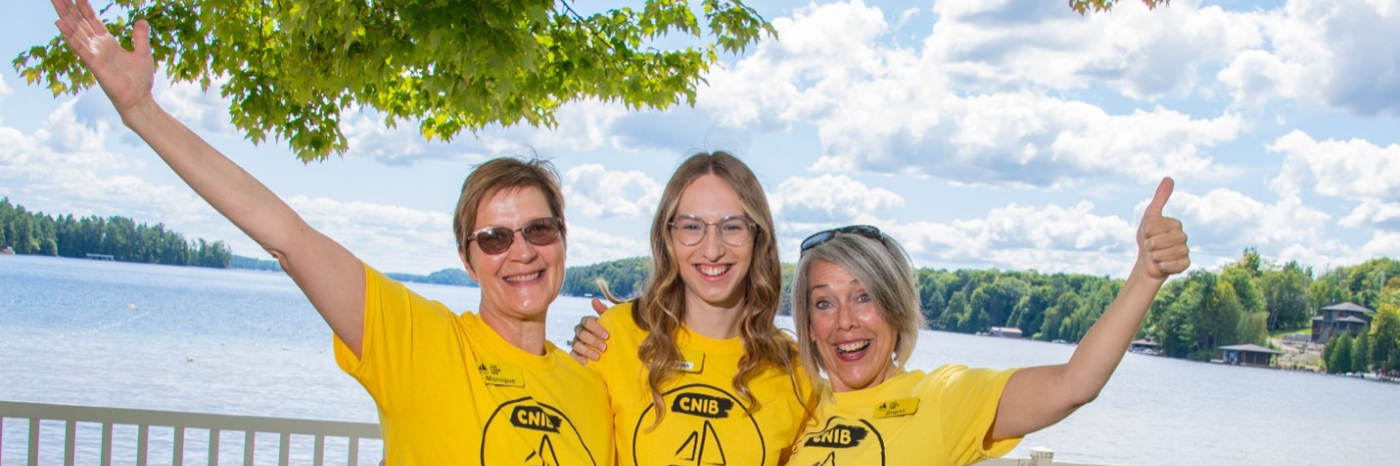 Monique Pilkington, Emma Van Dyk et Sherri Helsdingen se tiennent côte à côte au bord du lac Joe, vêtues de t-shirts jaunes « Lake Joe », les bras autour les unes des autres. Elles sourient à la caméra et lèvent le pouce.