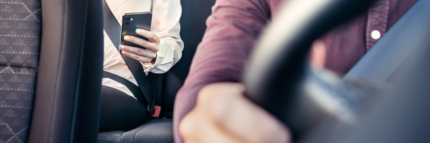 Un passager, attaché par sa ceinture de sécurité, est assis sur la banquette arrière d'une voiture de covoiturage, un téléphone intelligent à la main, tandis que le conducteur tient le volant depuis le siège avant.
