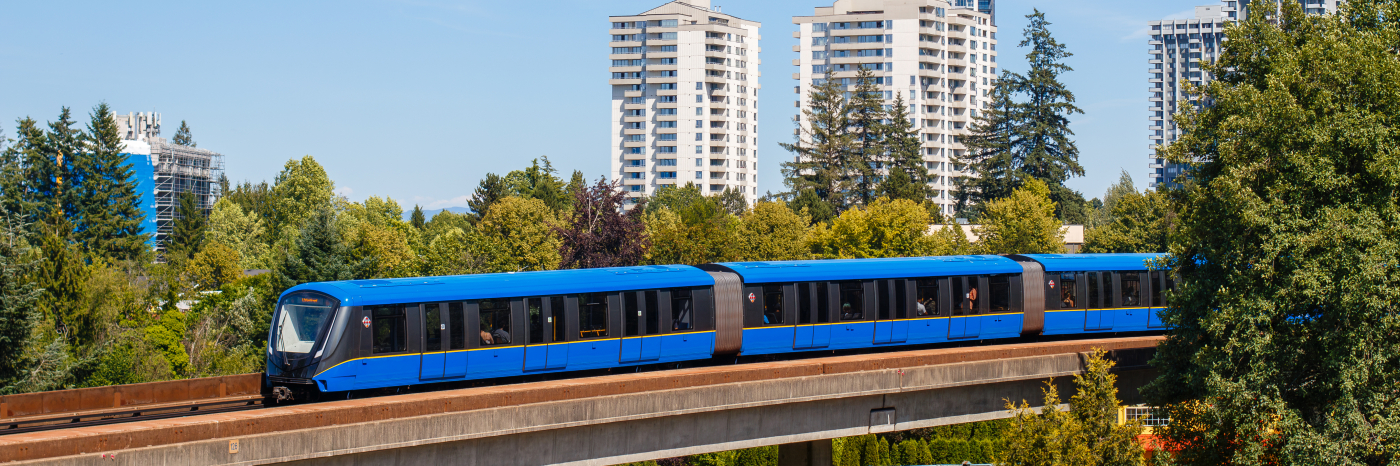 Une rame du SkyTrain de Vancouver circule sur une voie surélevée au-dessus d'un quartier arboré, parsemé d'immeubles résidentiels de grande hauteur, sous un ciel bleu limpide.