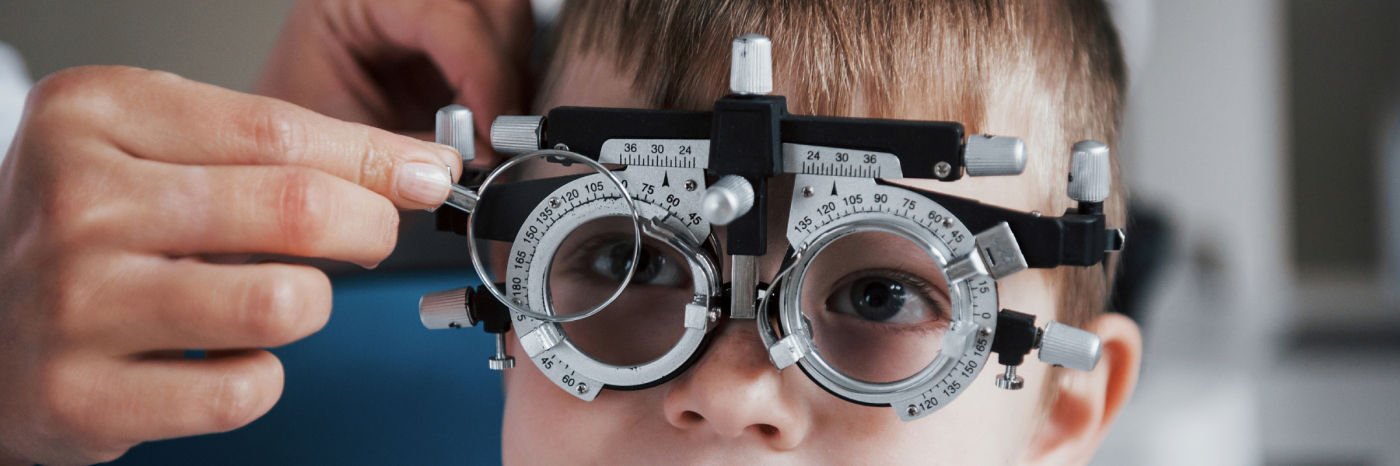 Un jeune enfant porte un phoropteur lors d'un examen de la vue, tandis qu'un professionnel de la vue ajuste l'un des verres.