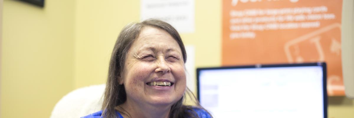 Woman sitting in a chair, smiling. Behind her is a computer screen and an orange poster on a yellow wall.