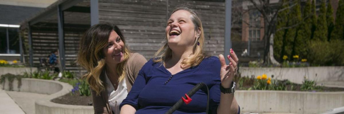 A brunette woman sits and smiles at a blond woman with a white cane, as both sit next to a sign for a fragrant garden.