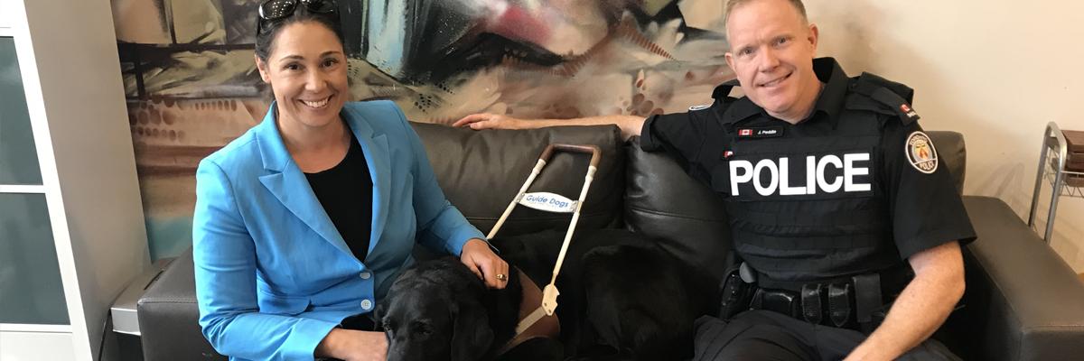 Victoria, her Guide Dog Alan, and a police officer sit on a couch.