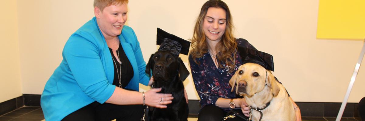 Ashley et Danika accompagnées de deux chiens Labrador/Golden Retriever croisés (un noir et un jaune) portant des chapeaux de graduation.