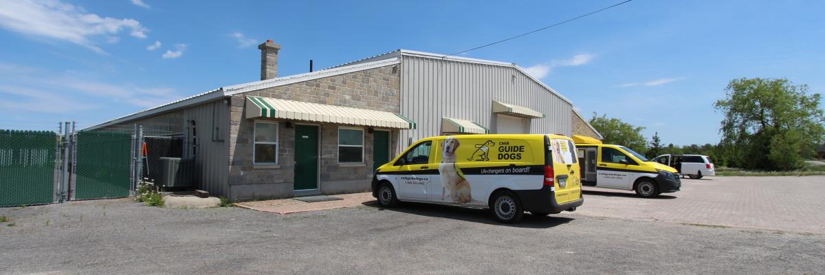 Exterior of Canine Campus in Carlton Place. Three CNIB Guide Dogs vans parked out front, trees and blue skies in the background.