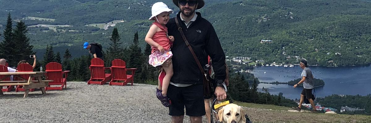 David avec sa fille dans un bras et de l’autre main, tenant dans son harnais son chien-guide Lilo, souriant pour la caméra, le Versant Sud du Mont Tremblant au Québec en arrière-plan