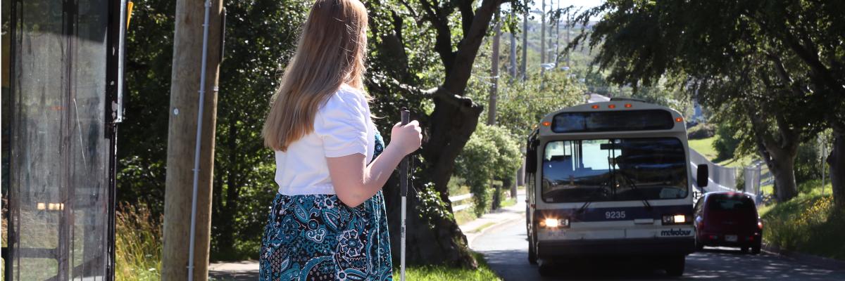 Une femme tenant une canne blanche se tenant devant un abribus attendant le bus qui est sur le point d’arriver.