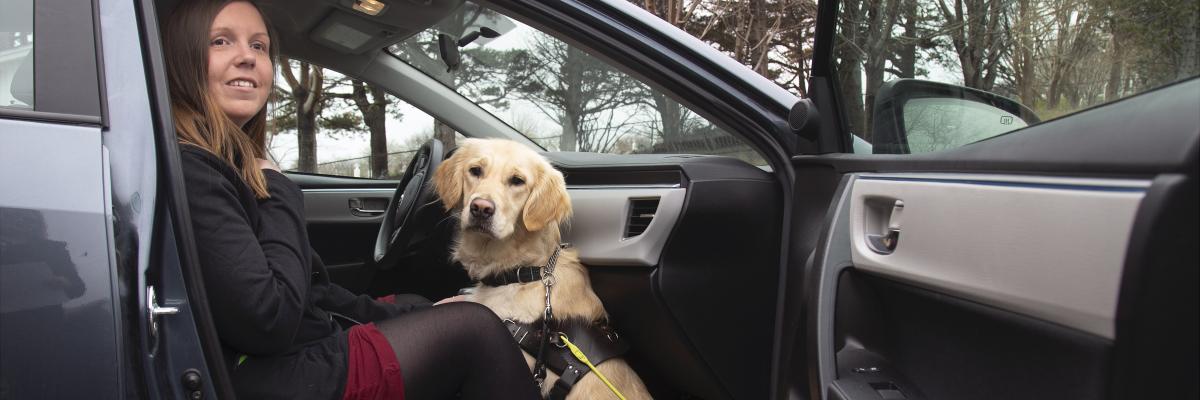 Kelly et de son chien-guide Maple, assis sur le siège côté passager d'une voiture la portière ouverte, souriant pour la caméra.