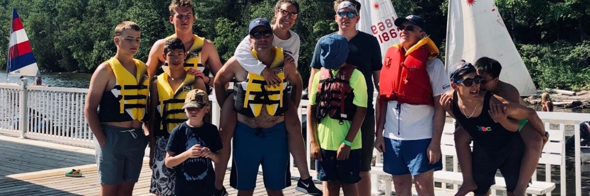 A group of people standing on the dock in shorts and life jackets. They are smiling and, in some cases, piggy-backing others.