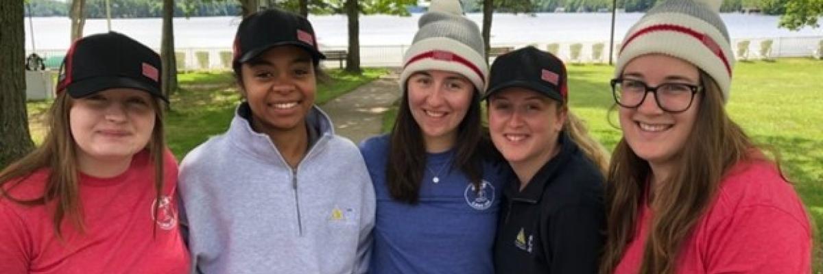 : Five young women (CNIB Lake Joe staff) pose for a photo together. They are wearing CNIB Lake Joe t-shirts and hats.