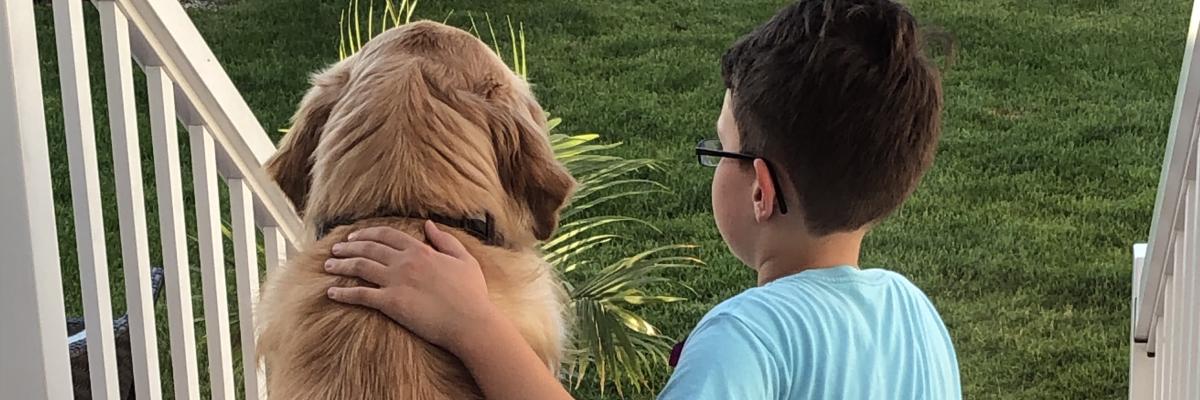 The backs of Austin and his CNIB Buddy Dog Dickson, a golden retriever, sitting on an outdoor backstep looking out; Austin is petting Dickson’s back.