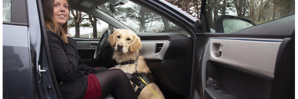 A woman sitting in the front passenger seat of a taxi with her golden retriever guide dog sitting on the floor at her feet with the car door open.