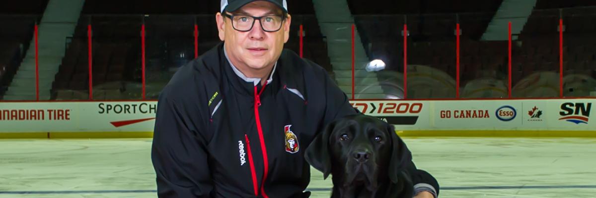 Mark Kelly à genoux à côté de Rookie, un futur chien-guide d'INCA portant un maillot des Sénateurs d'Ottawa, avec son bras autour de Rookie tandis qu'ils sourient tous les deux pour la caméra. Ils se trouvent sur un tapis rouge posé sur la glace du Centre Canadian Tire, domicile des Sénateurs d'Ottawa.