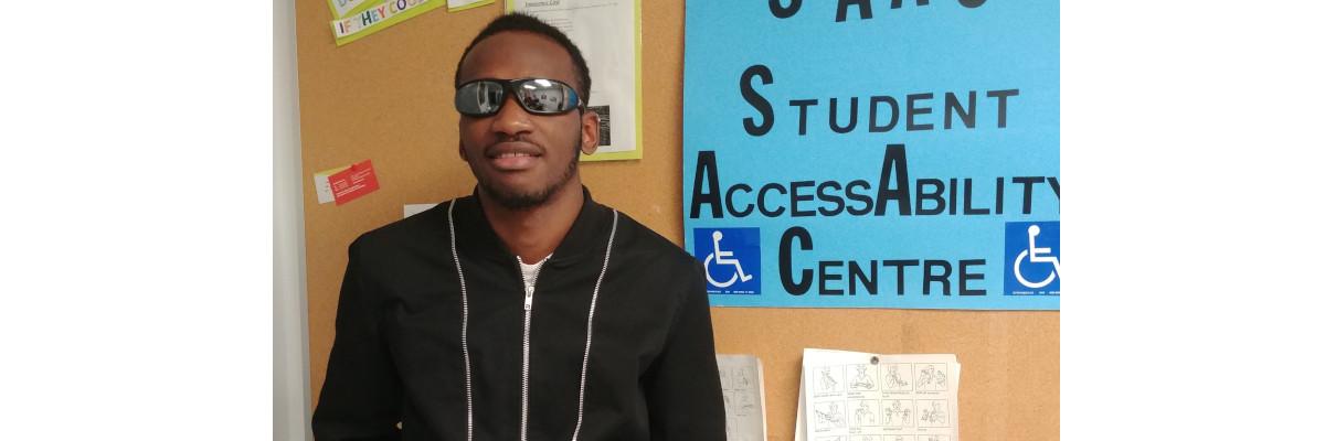  Eitel Houedakor smiling and standing in front of a bulletin board. He is wearing sunglasses and a hoodie.