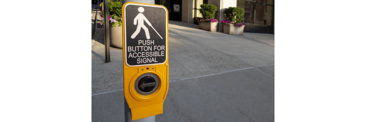 A yellow/black Accessible Pedestrian Signal pushbutton mounted to a pole.