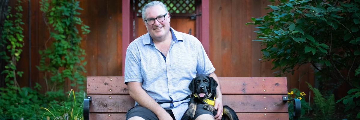 Christopher Adams sitting on a bench with a very young Percy sitting beside him wearing his bright yellow Future CNIB Guide Dogs vest.