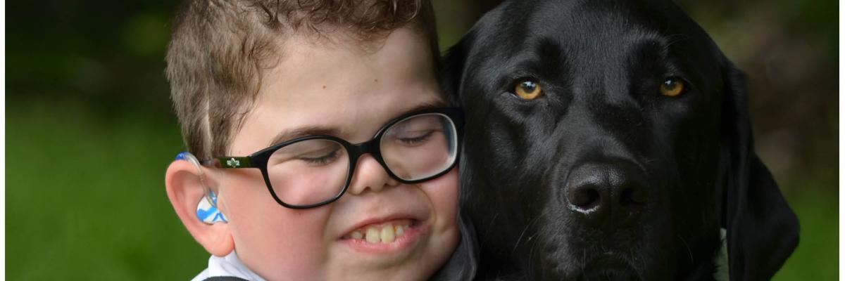 Kaiden has his eyes closed and is leaning into Flinn and giving him a big hug;  Flinn, a black lab, is sitting looking at the camera.