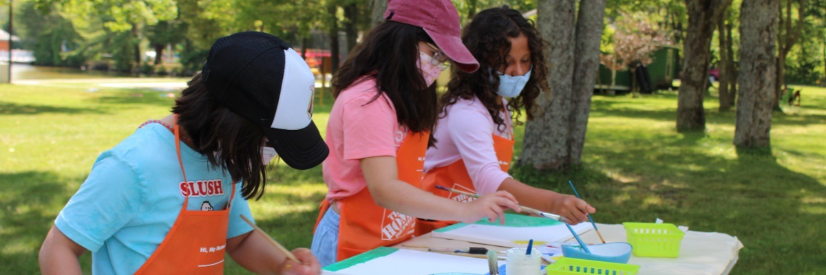 Three girls painting at a crafts table set up outside on the lawn.