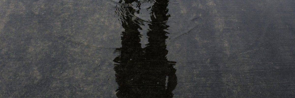 Un jeune homme se tient debout et regarde au-dessus d'une flaque d'eau. Ses chaussures de course apparaissent en haut de l'image. Dans la flaque d'eau apparaît un reflet délicat de sa silhouette.