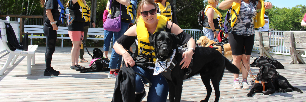  group of guide dog handlers wearing lifejackets stand on the dock at CNIB Lake Joe with their guide dogs. Taylor crouches and poses in the foreground next to her guide dog, Wallace.