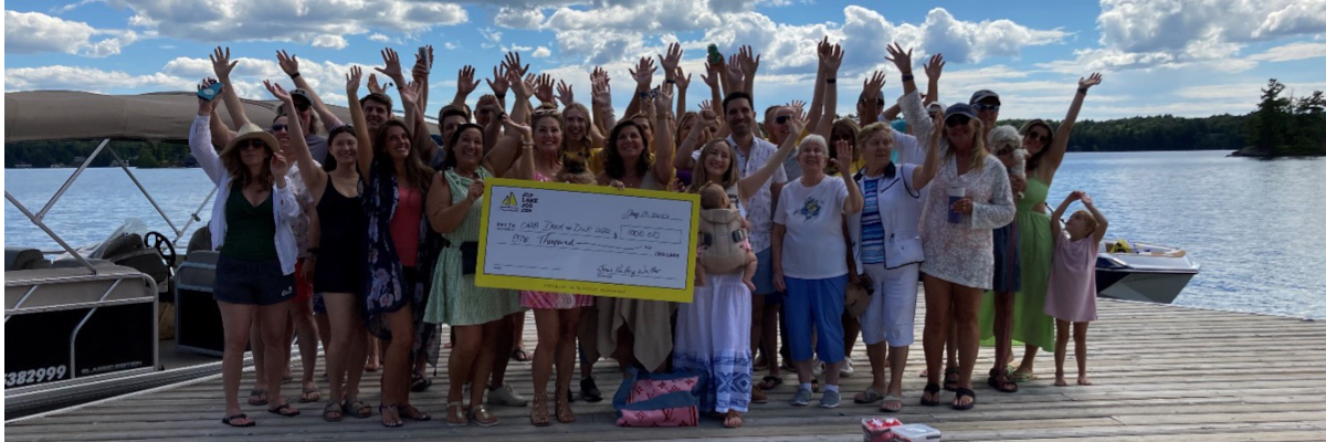 Un groupe d'invités debout sur un quai au bord de l'eau et salue avec enthousiasme, en montrant leur don d’un chèque géant de 1 000 $. 