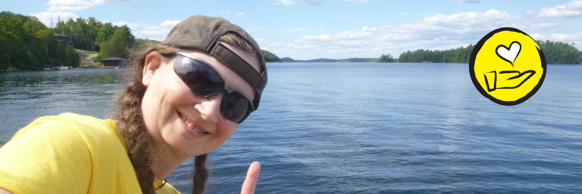 Jessica Bailey, poses at the Lake Joe waterfront. She does the American Sign Language sign for "I Love you". An icon of a hand with a heart floating above it appears in the top right hand corner. 