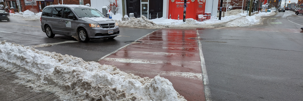 A pile of plowed snow blocks the sidewalk at a busy crosswalk